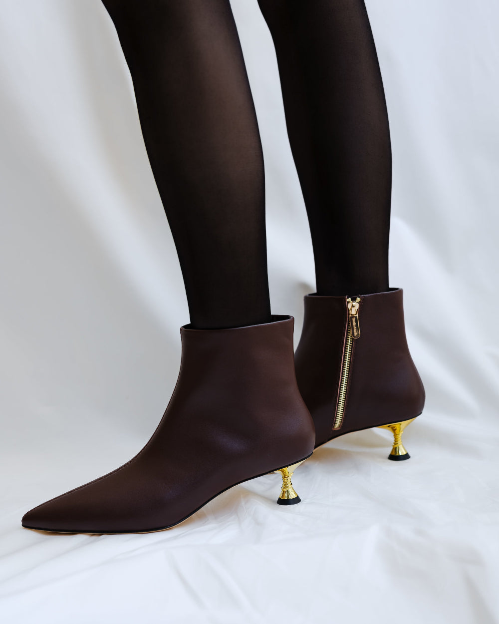 Brown ankle boots with gold zippers and thin legs wearing black tights on a white background
