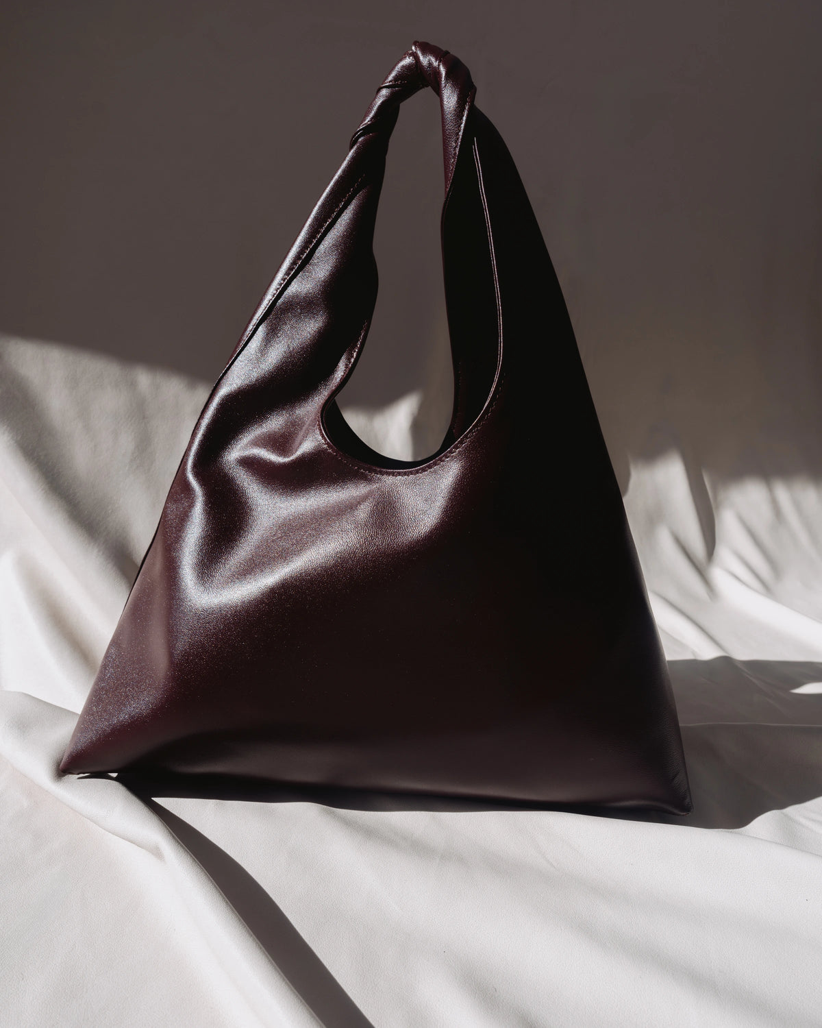 A slouchy, triangular leather shoulder bag in deep burgundy, resting on draped white fabric with dramatic, directional sunlight casting soft shadows.