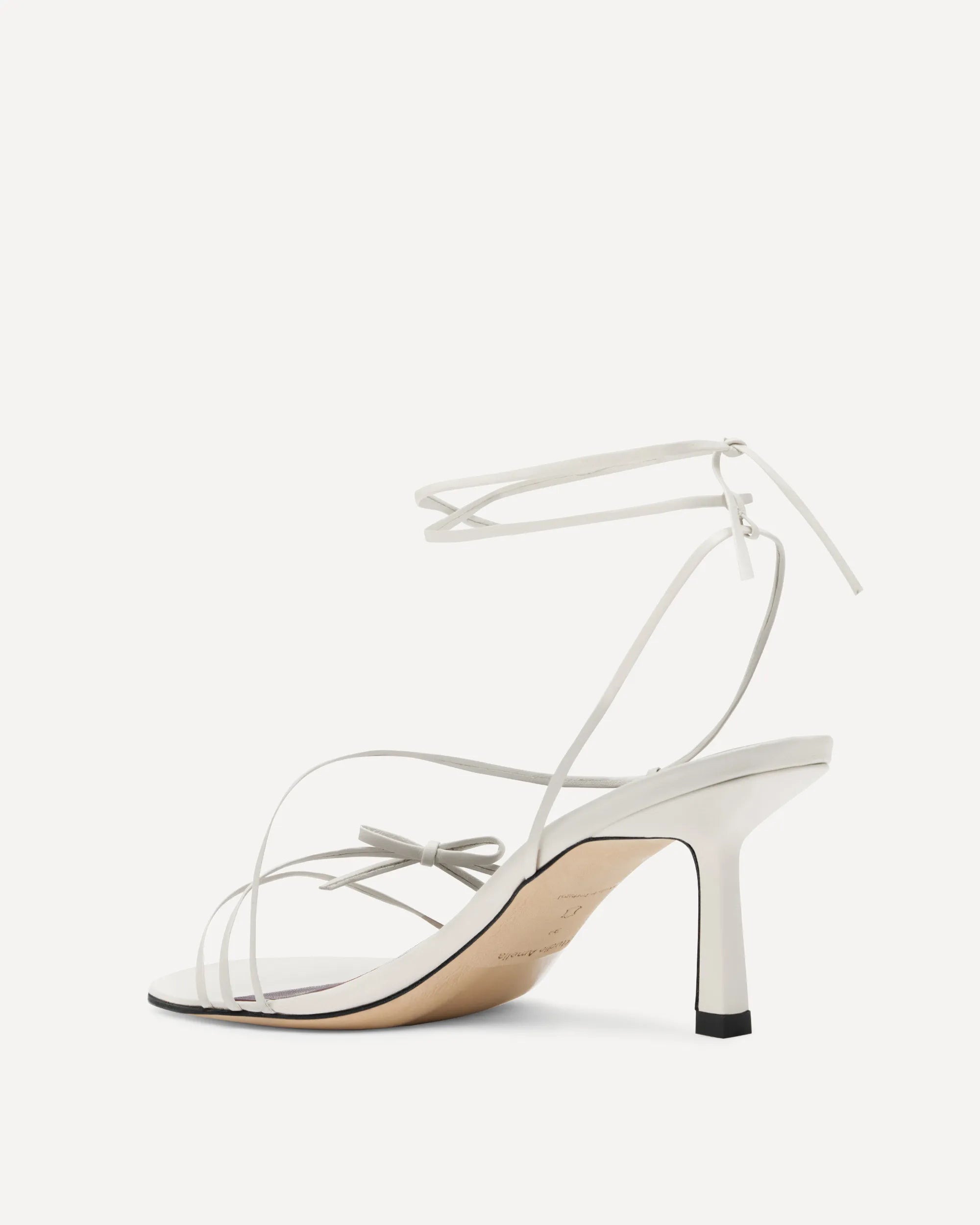 A single white leather high-heeled sandal with thin wraparound ankle straps, a small bow detail on the instep, and a slim stiletto heel, shown from a side profile against a light off-white background.