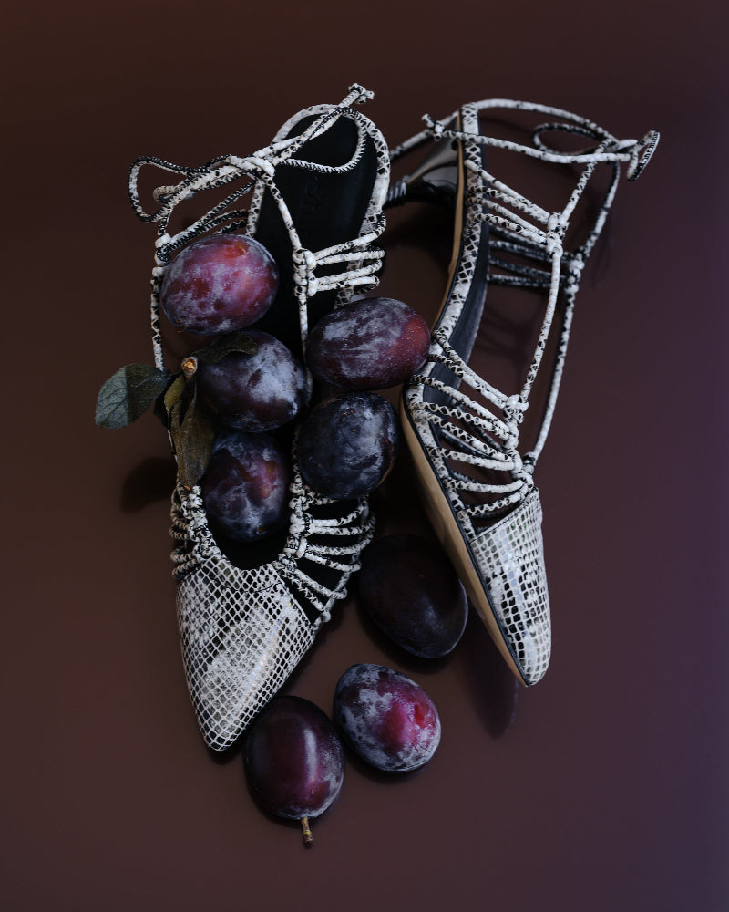 Pair of snake-skin high heels next to plums on a brown background