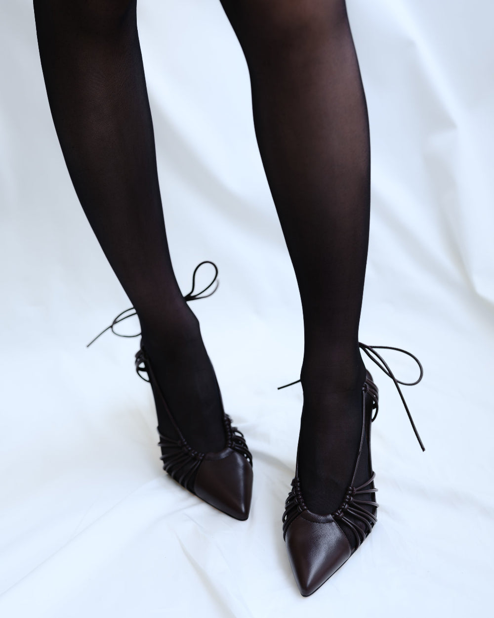 Black high-heeled shoes with ruched details worn with black tights on a white background