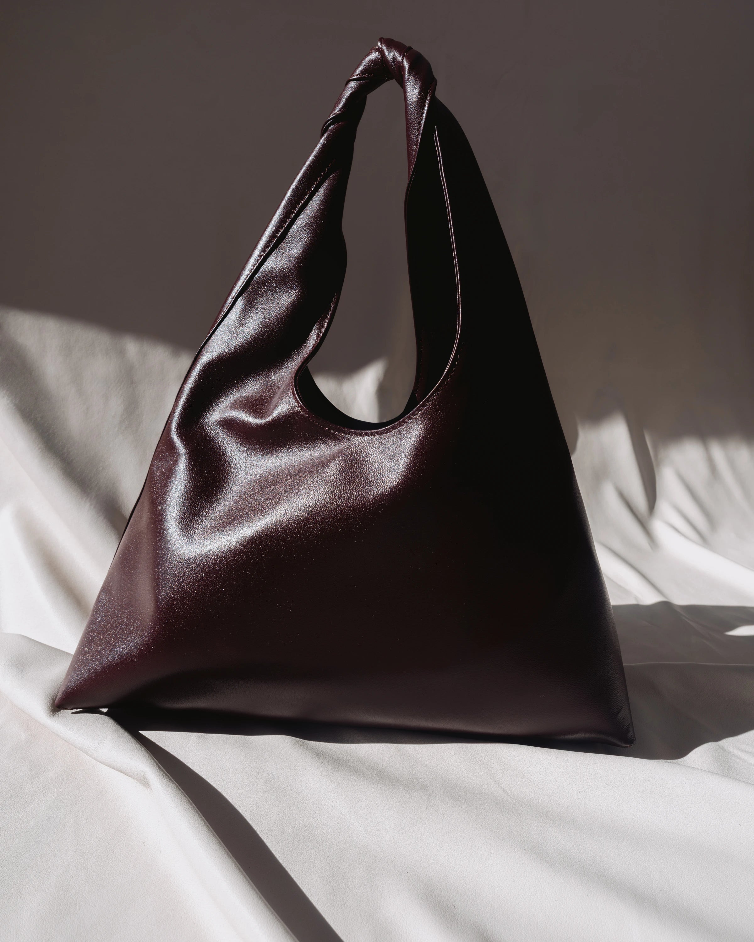 A slouchy, triangular leather shoulder bag in deep burgundy, resting on draped white fabric with dramatic, directional sunlight casting soft shadows.