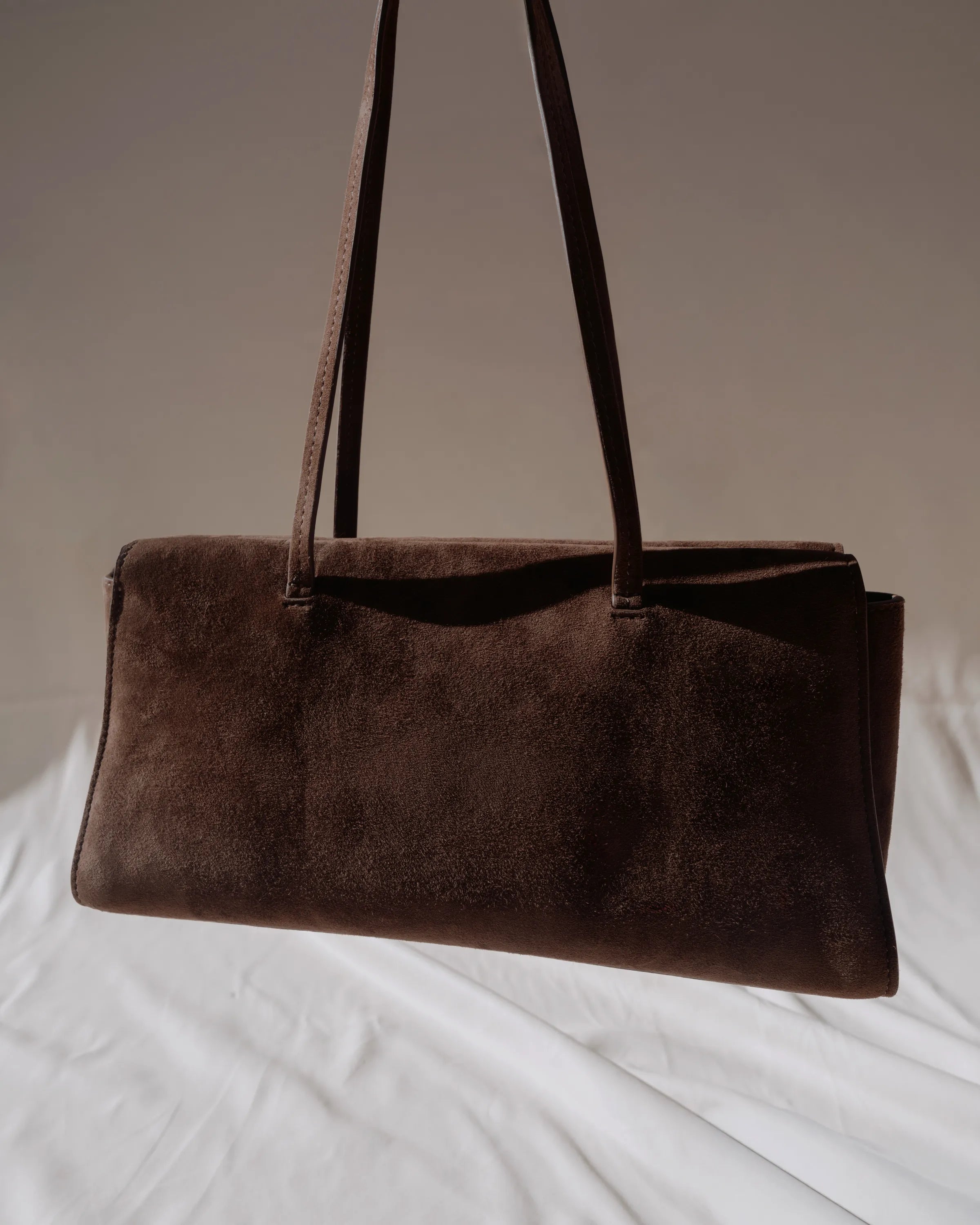 A dark brown suede shoulder bag with a long strap, resting on white fabric against a neutral beige background.