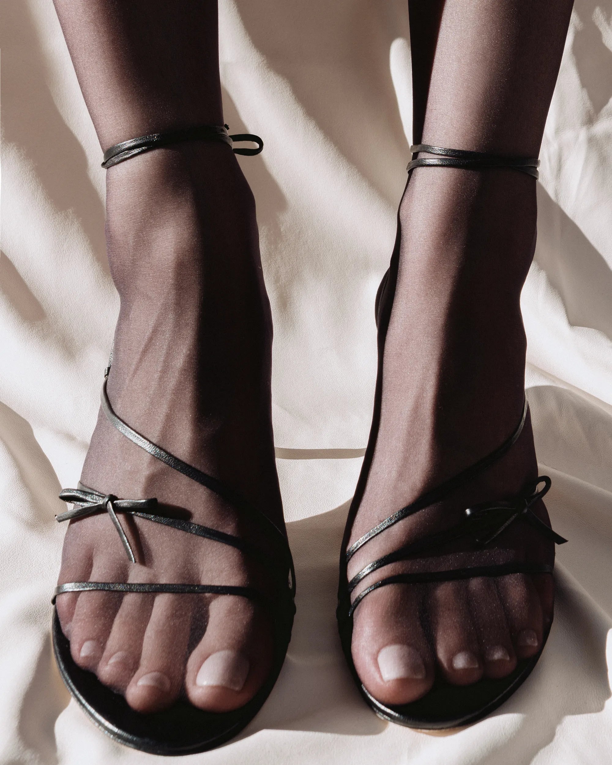 Black strappy flat sandals with ankle ties worn with sheer black tights, shown on feet against draped cream fabric.
