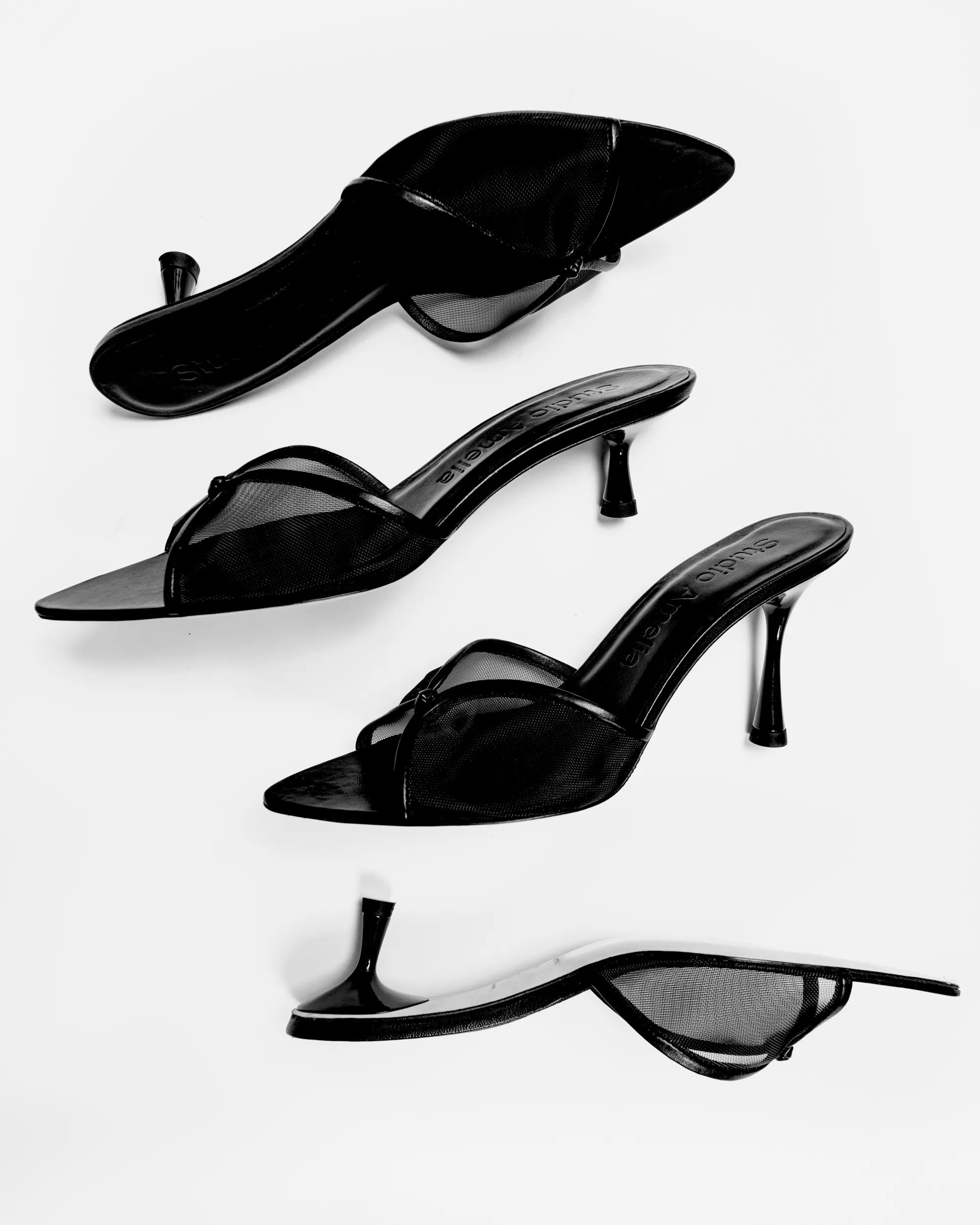 Four black high-heeled mule sandals with pointed toes and thin straps, arranged diagonally on a light gray background.