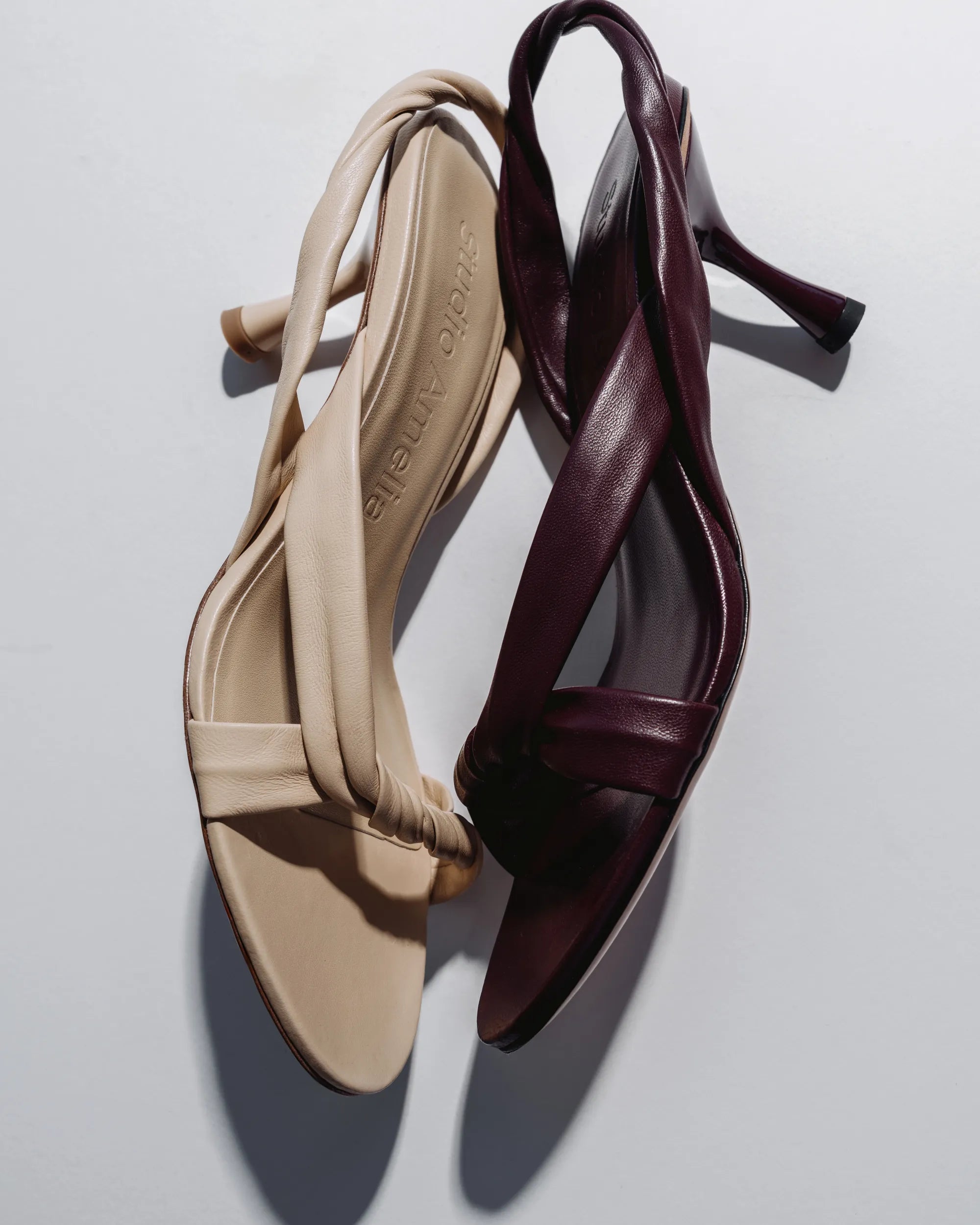 Two elegant slingback high-heeled sandals with crisscross straps, shown overlapping on a light gray background. One sandal is beige and the other is deep burgundy.