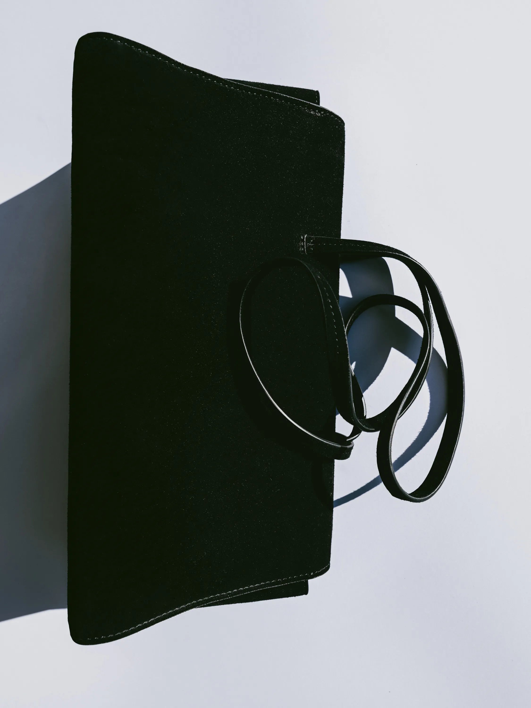A black envelope-style clutch bag with a long thin shoulder strap, photographed on a light gray background with a strong diagonal shadow.