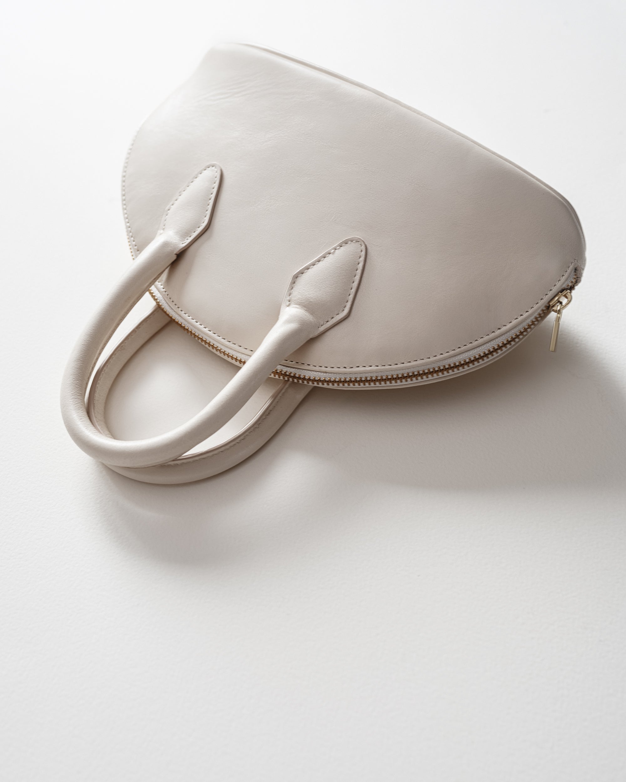 A light beige leather handbag with a unique crescent shape, dual top handles, and a gold zipper, photographed on a clean white background.